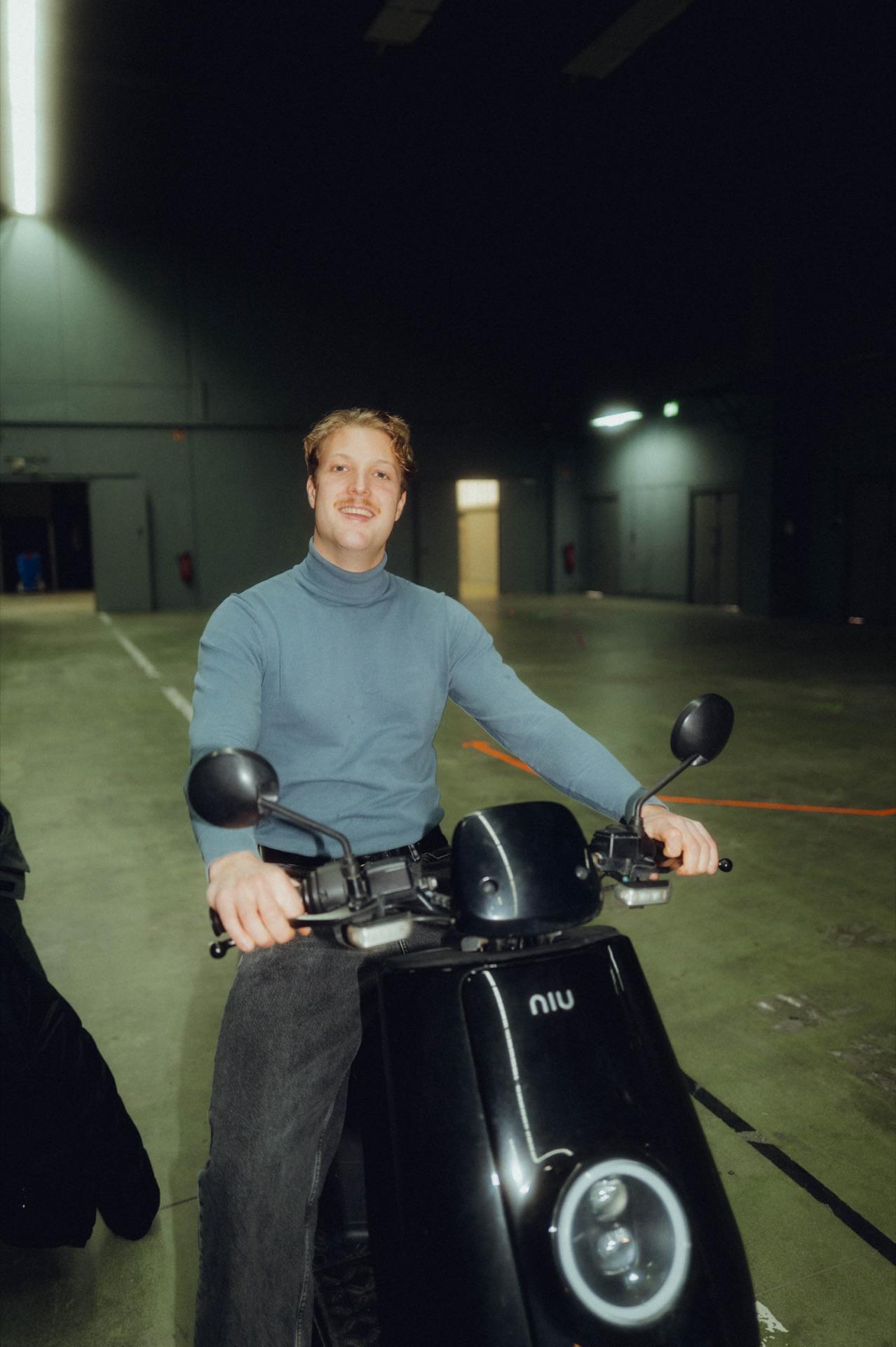 Crew-Mitglied im Rollkragen sitzt lachend auf einem Motorroller in der Halle