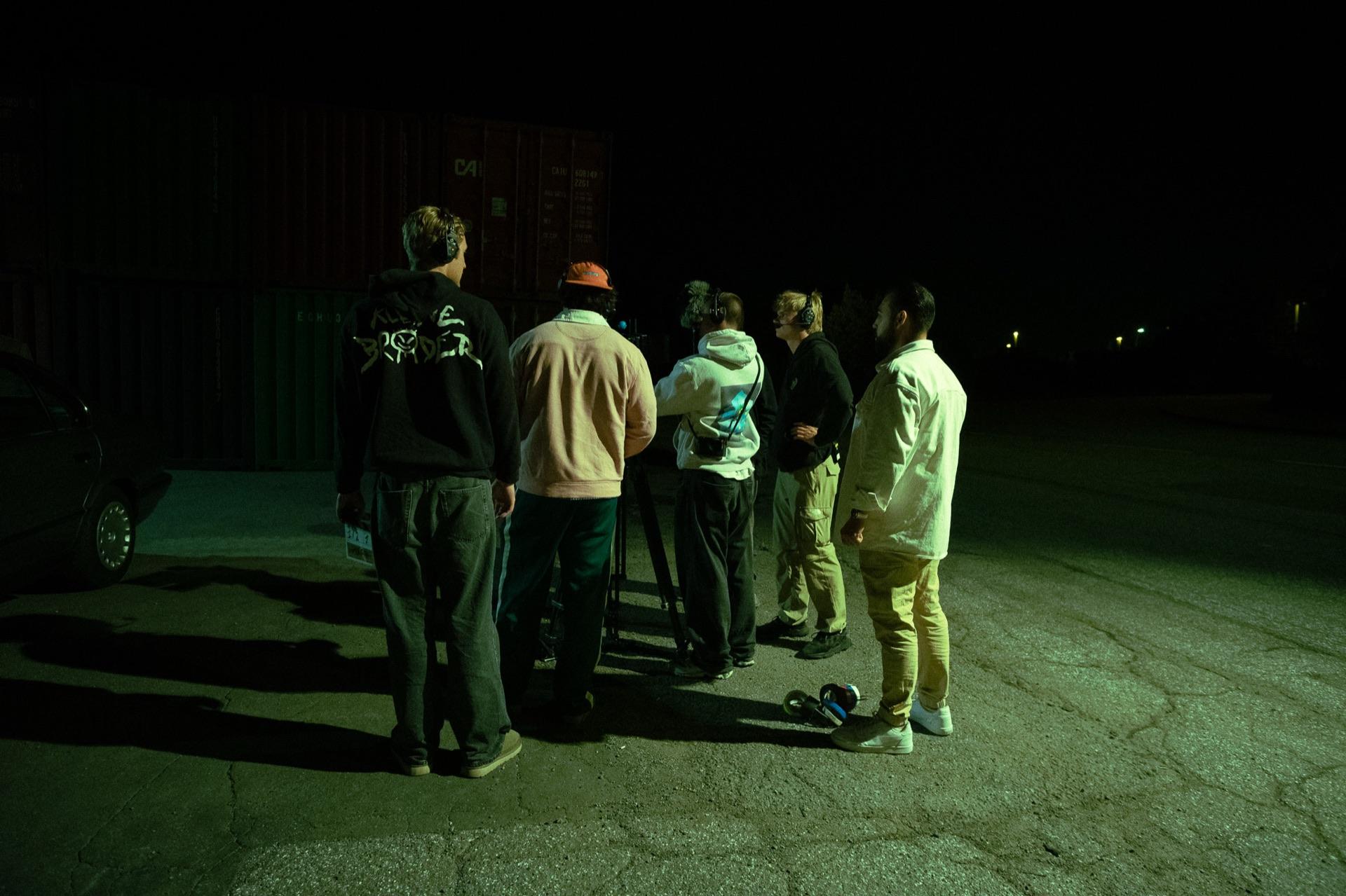Behind the Scenes: das Team steht nachts vor der Container-Wand zusammen, eine Filmlampe leuchtet den Platz aus