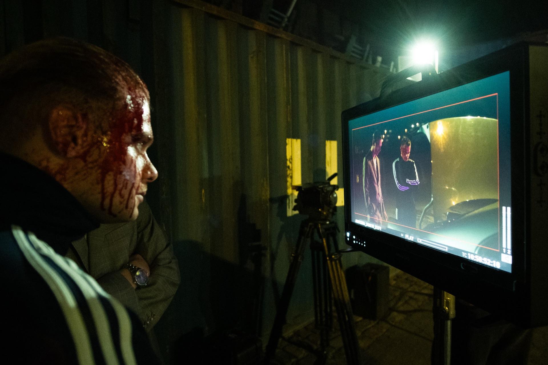 Behind the Scenes: der Darsteller mit Blut-Make-up blickt im Profil auf den Monitor neben der Kamera