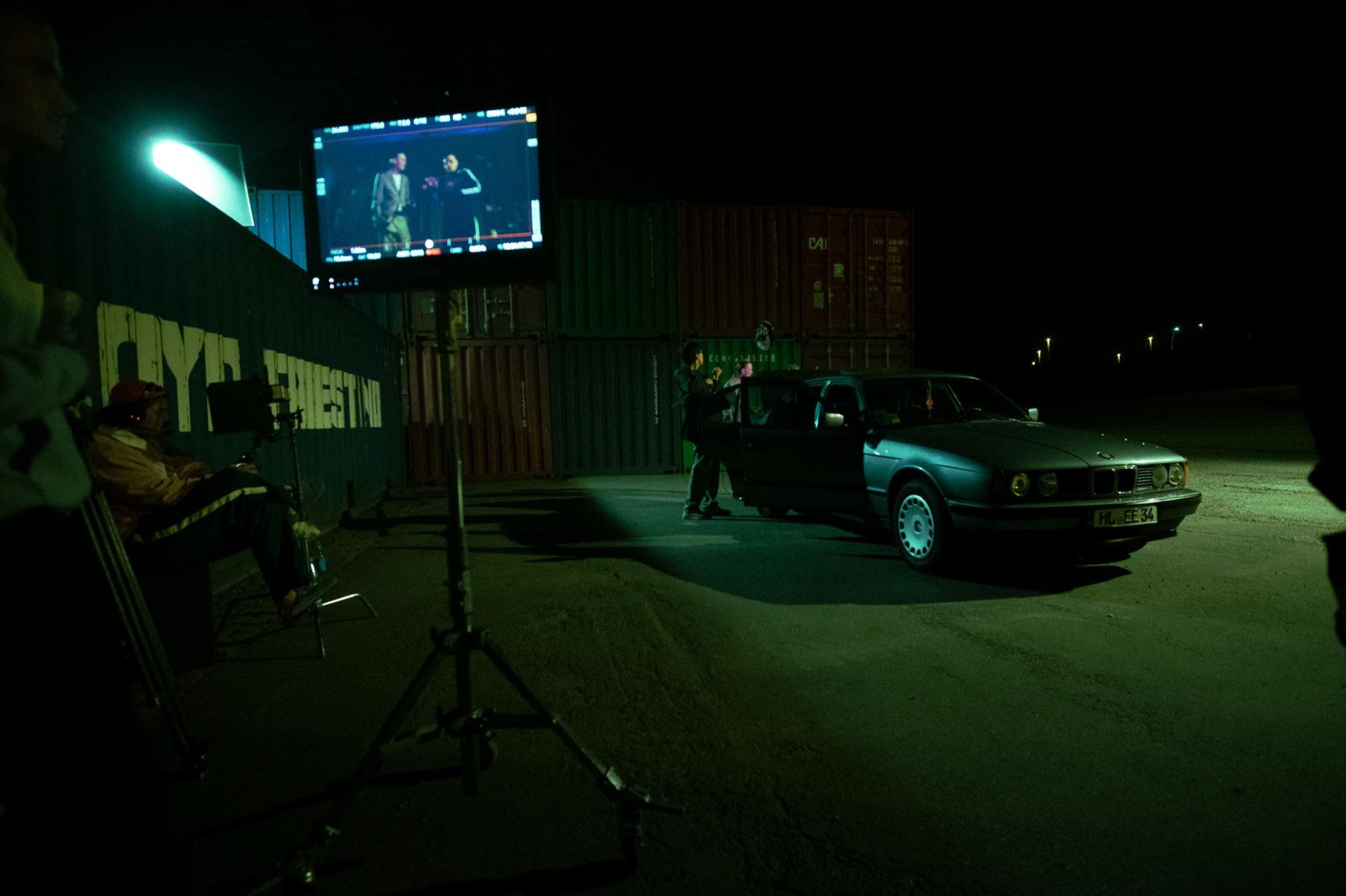 Behind the Scenes: Set-Überblick mit Monitor auf dem Stativ und der Crew am alten BMW vor den Containern