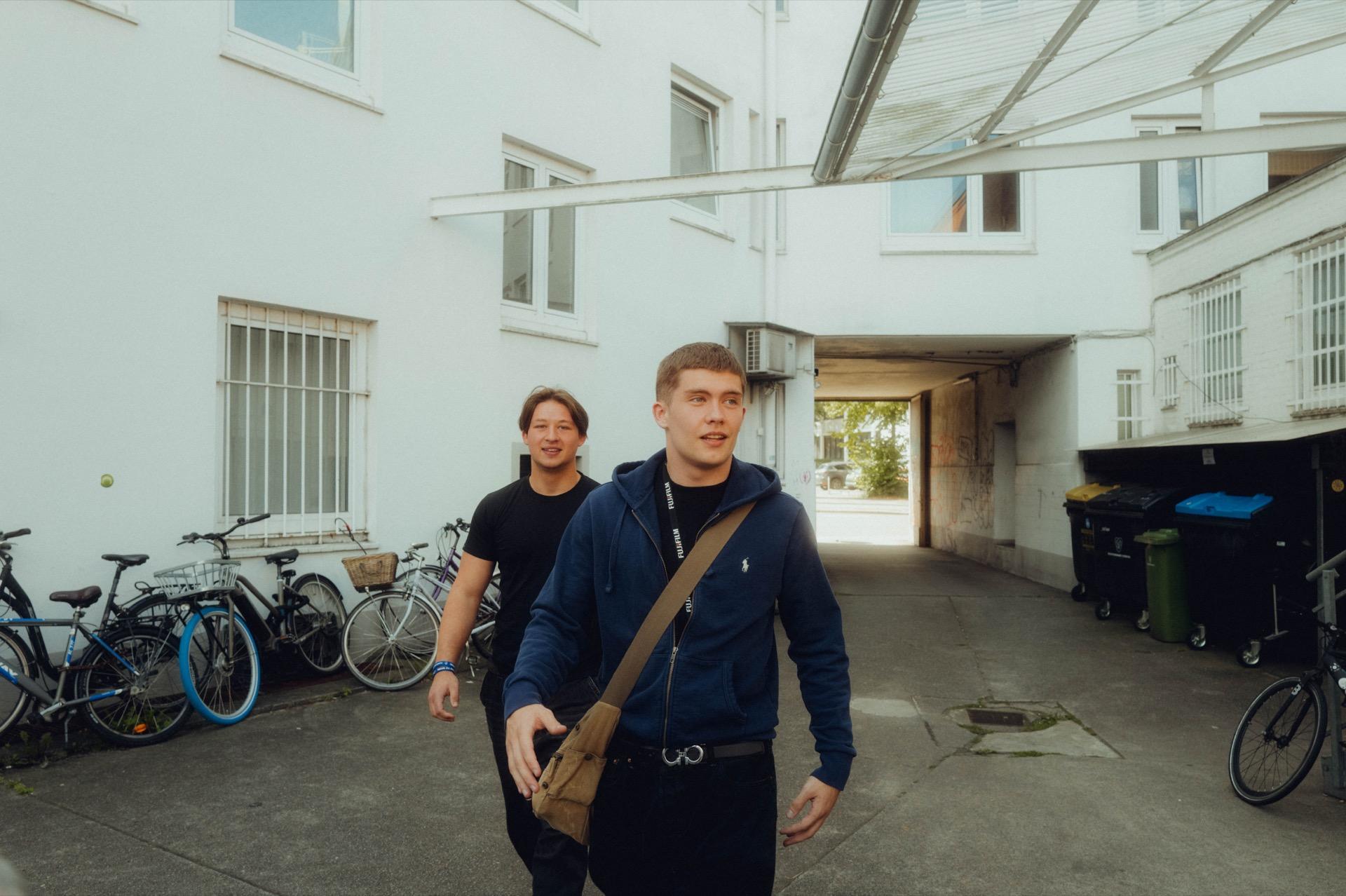 Behind the Scenes: zwei junge Männer gehen durch einen Hamburger Hinterhof, links eine Reihe abgestellter Fahrräder, im Hintergrund eine Tordurchfahrt