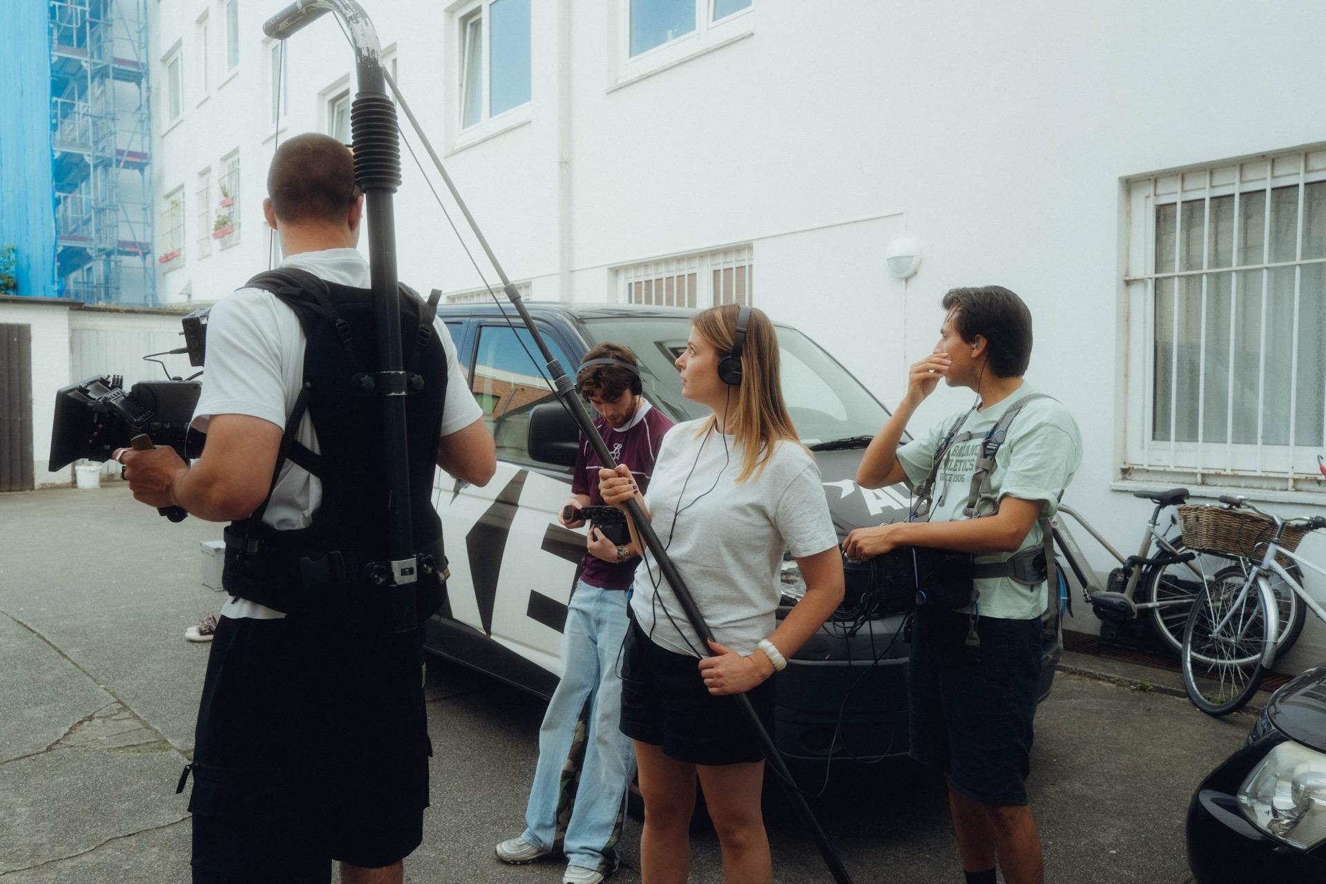 Behind the Scenes: Kameramann von hinten neben einer Tonangel, dahinter filmt die Crew eine Szene an einem silbernen Auto
