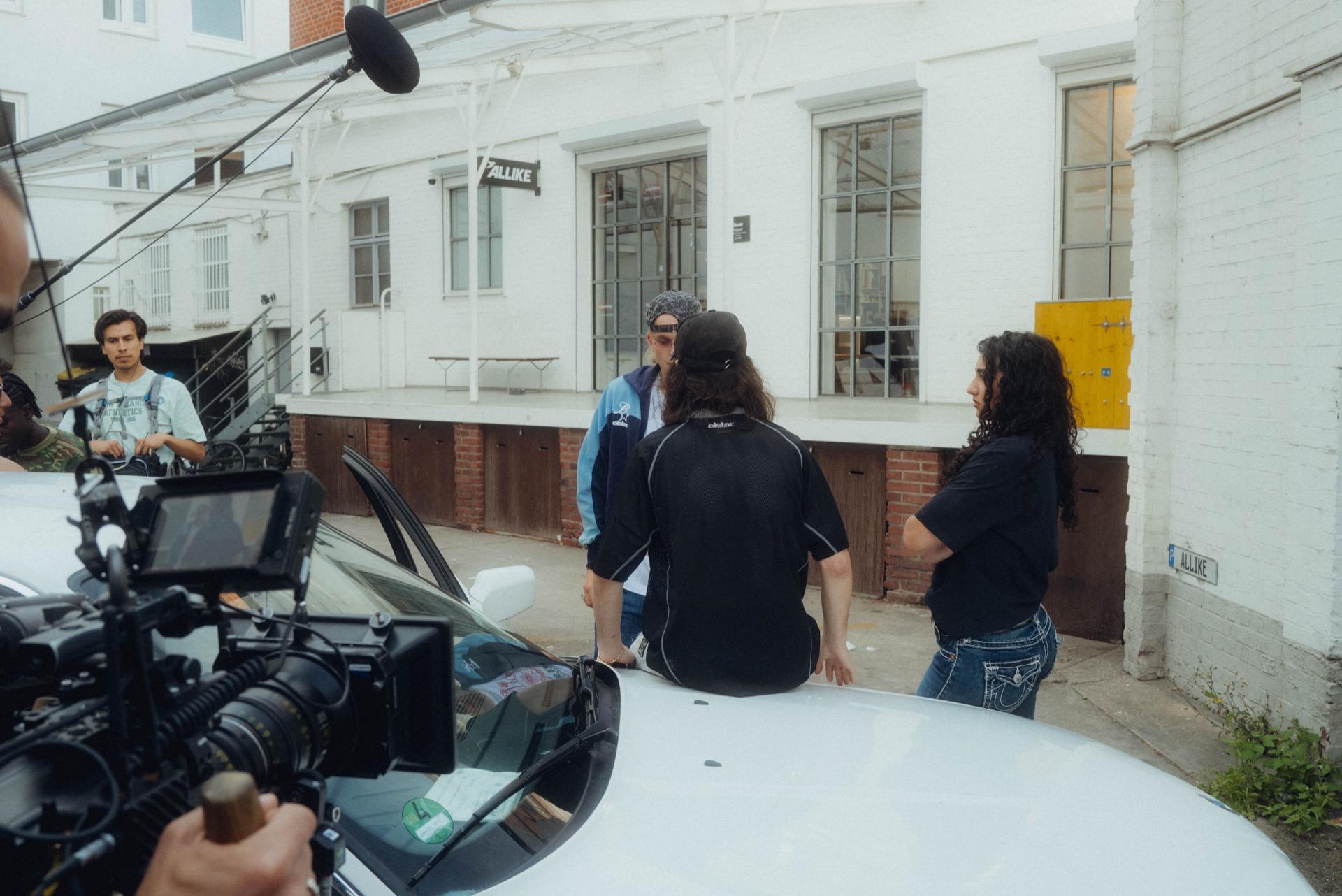 Behind the Scenes: eine Filmkamera auf einem Rig im Vordergrund, dahinter dreht die Crew vor einer weißen Fabrikfassade mit der Aufschrift ALIKE