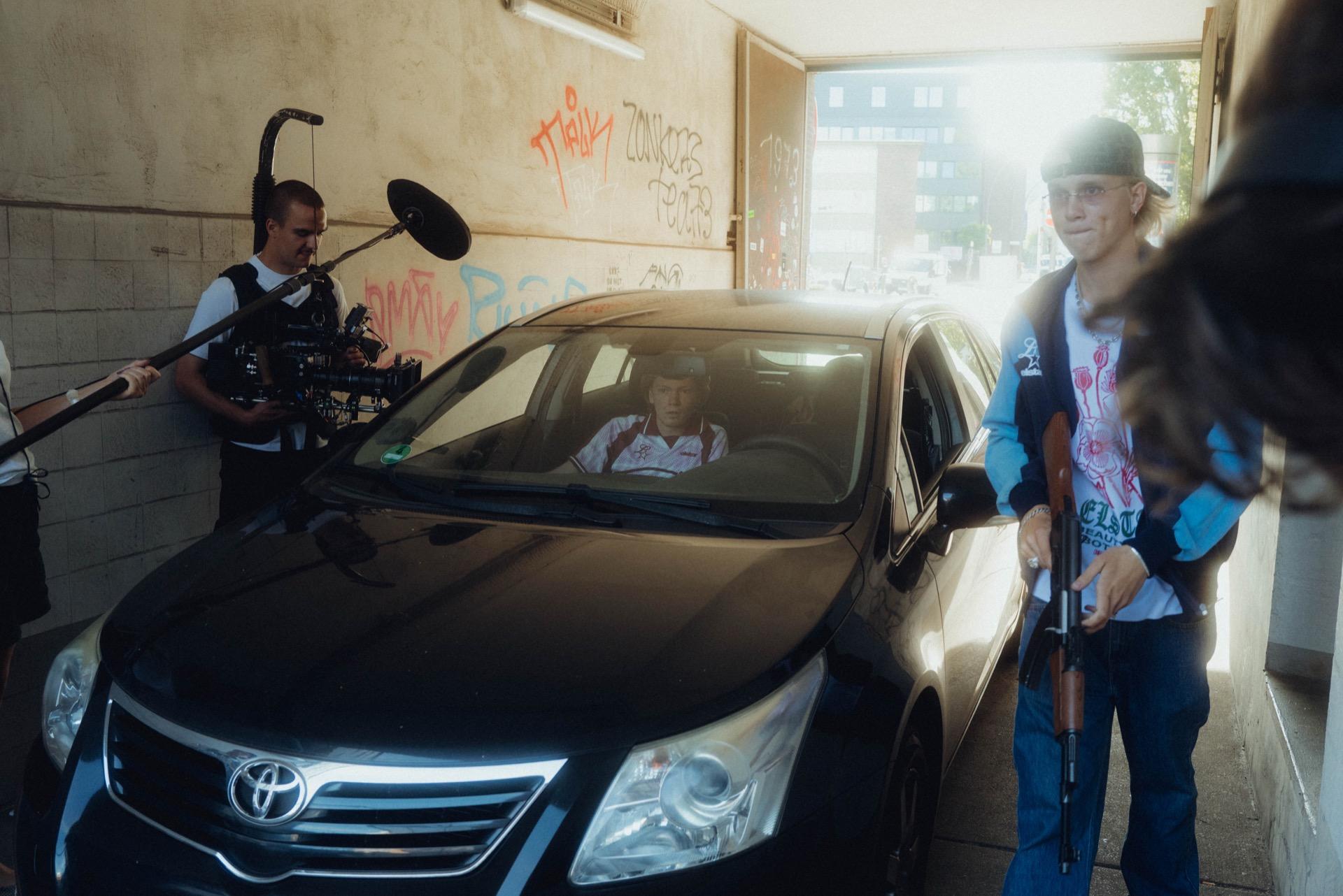 Behind the Scenes: in einer Tordurchfahrt im Gegenlicht filmt die Crew den schwarzen Toyota, rechts steht ein junger Mann in heller Jacke mit einem Requisit