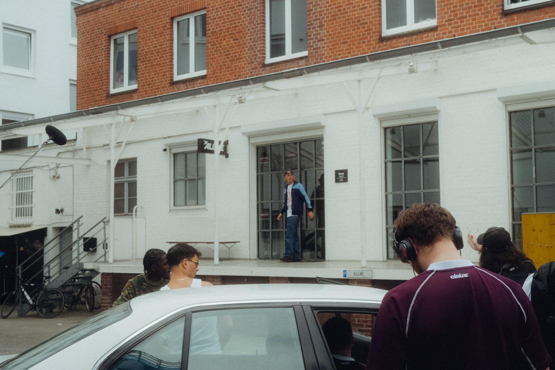 Behind the Scenes: weiter Blick auf das Set, ein Schauspieler steht in der gläsernen Eingangstür der weißen Halle, davor die Crew und ein weißes Auto