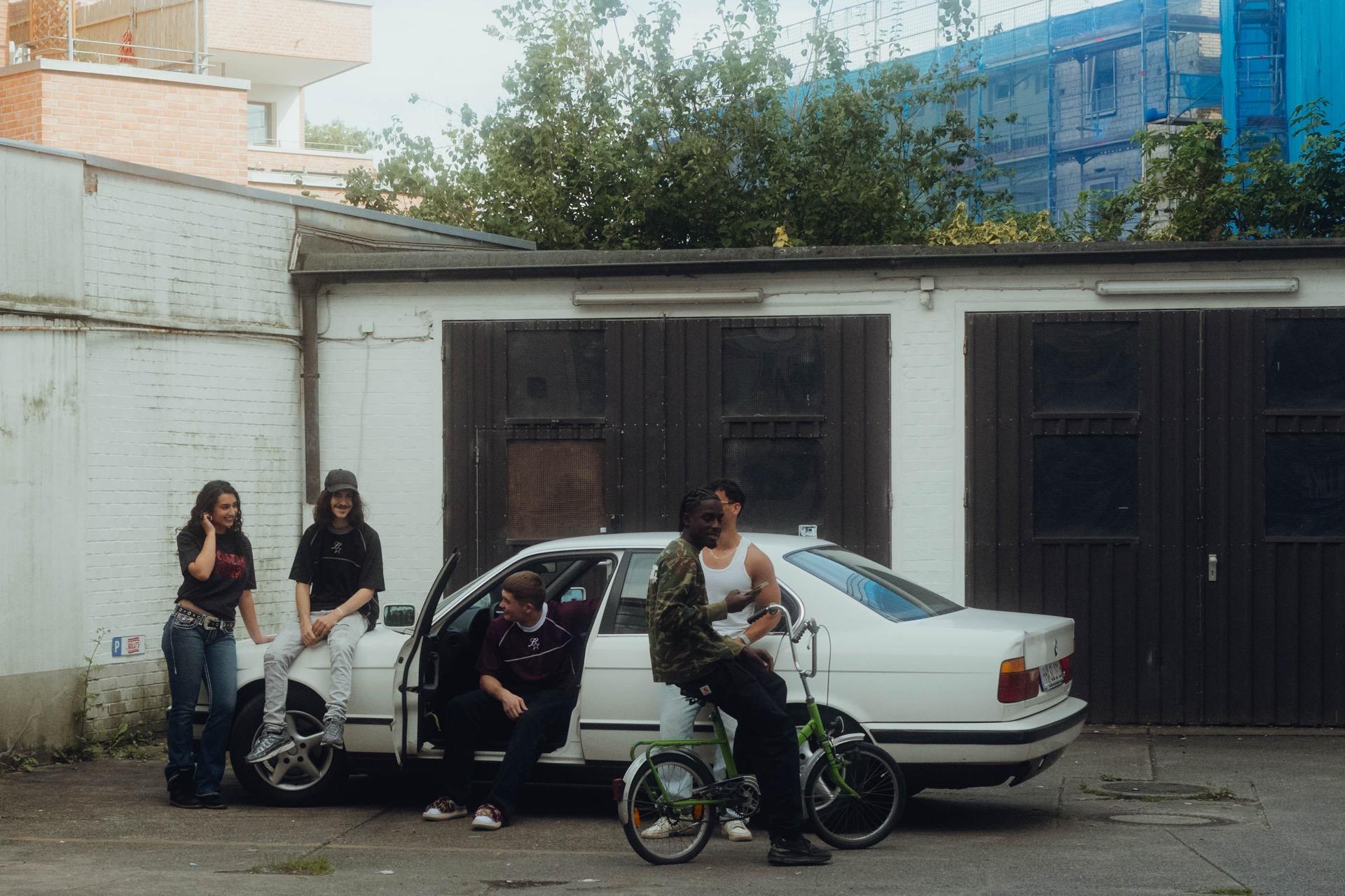 Behind the Scenes: mehrere Cast-Mitglieder lehnen an einem alten weißen BMW vor Garagentoren, einer sitzt auf einem klappbaren BMX-Rad