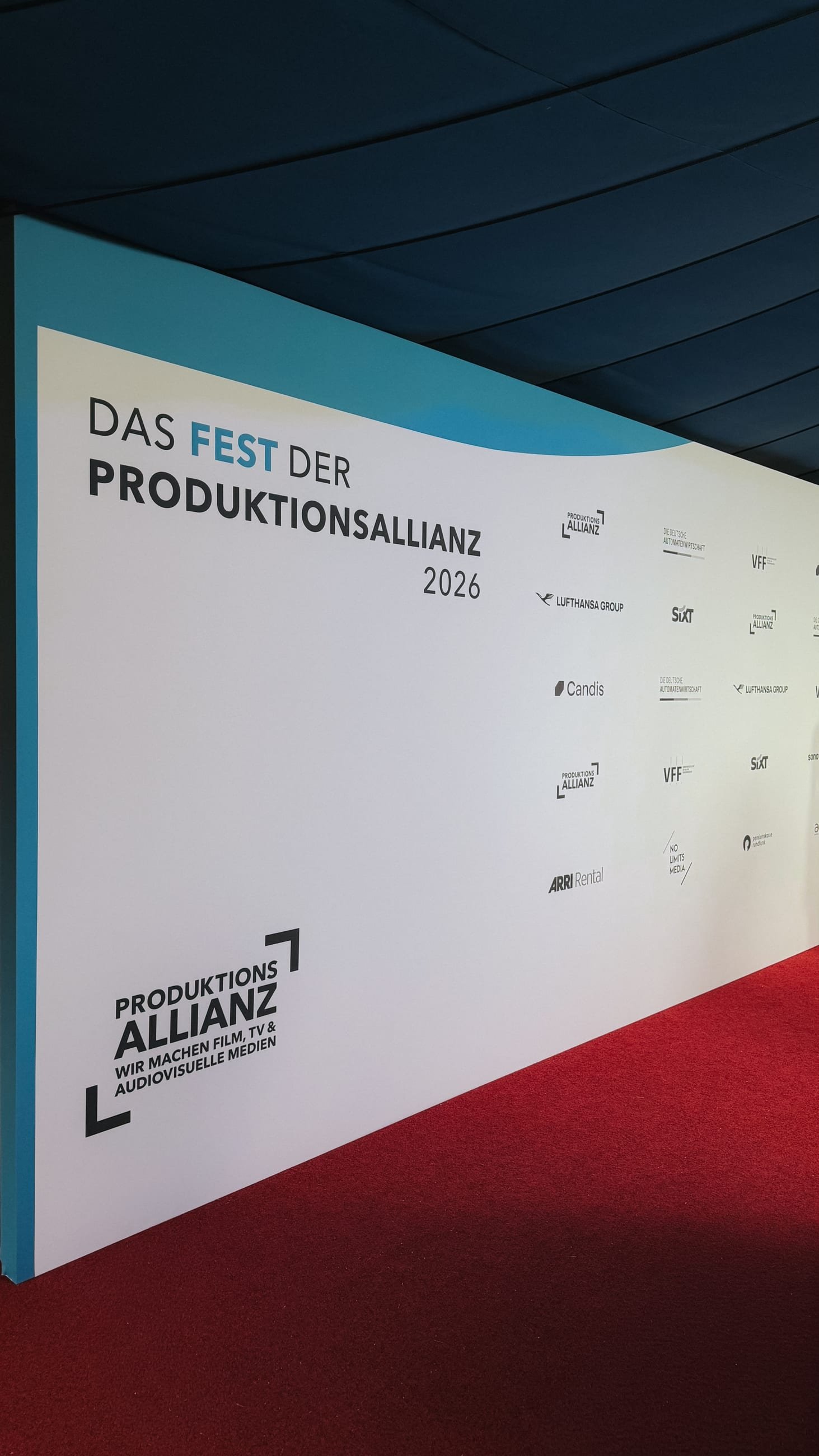 Fotowand mit dem Schriftzug „Das Fest der Produktionsallianz 2026“ und den Logos der Partner, davor roter Teppich, darüber die blaue Zeltdecke