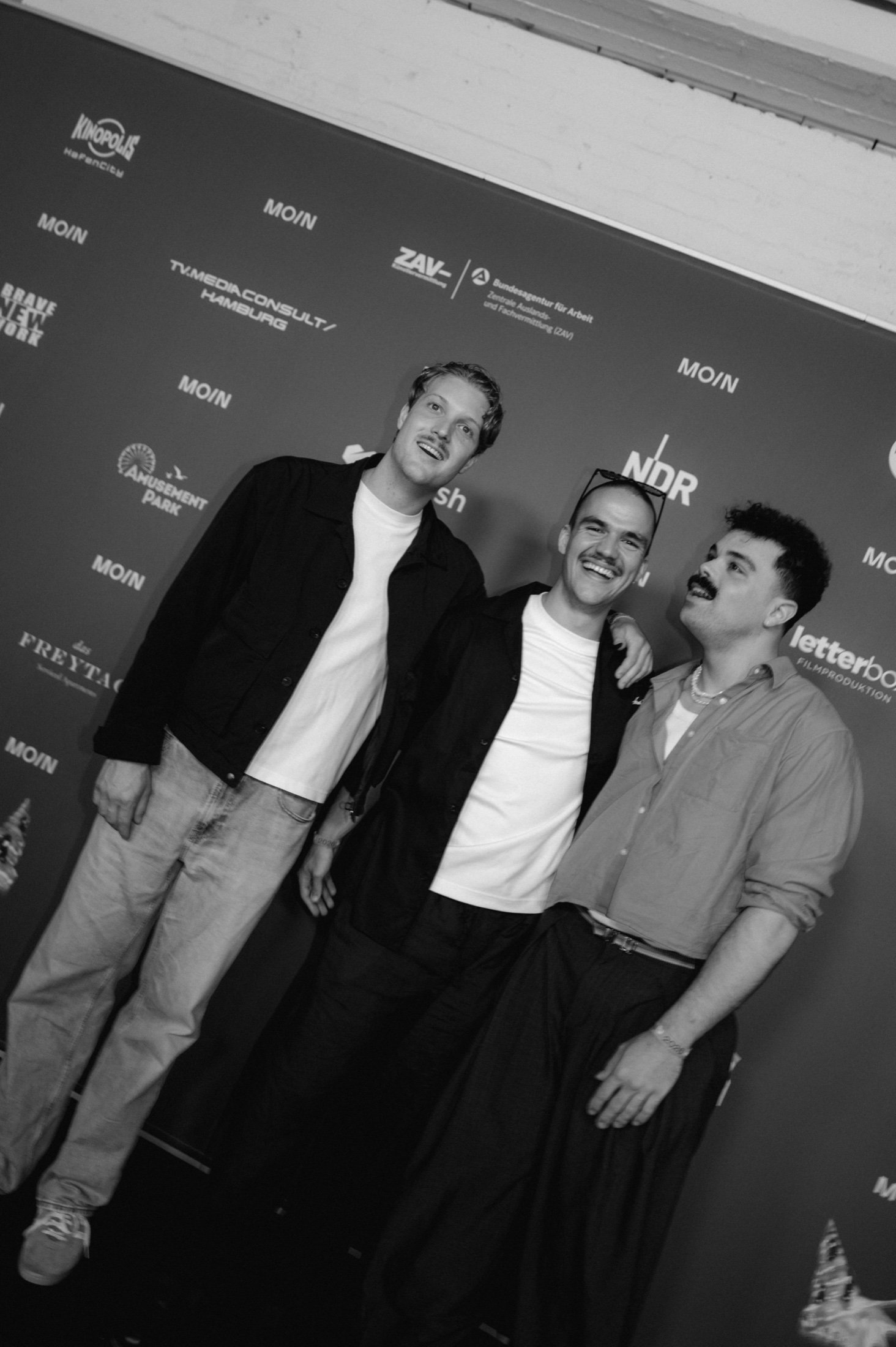 Jakob, Johannes und Lino von [k]eine produktion lachen vor der MOIN Fotowand mit den Logos von NDR, ZAV und weiteren Partnern