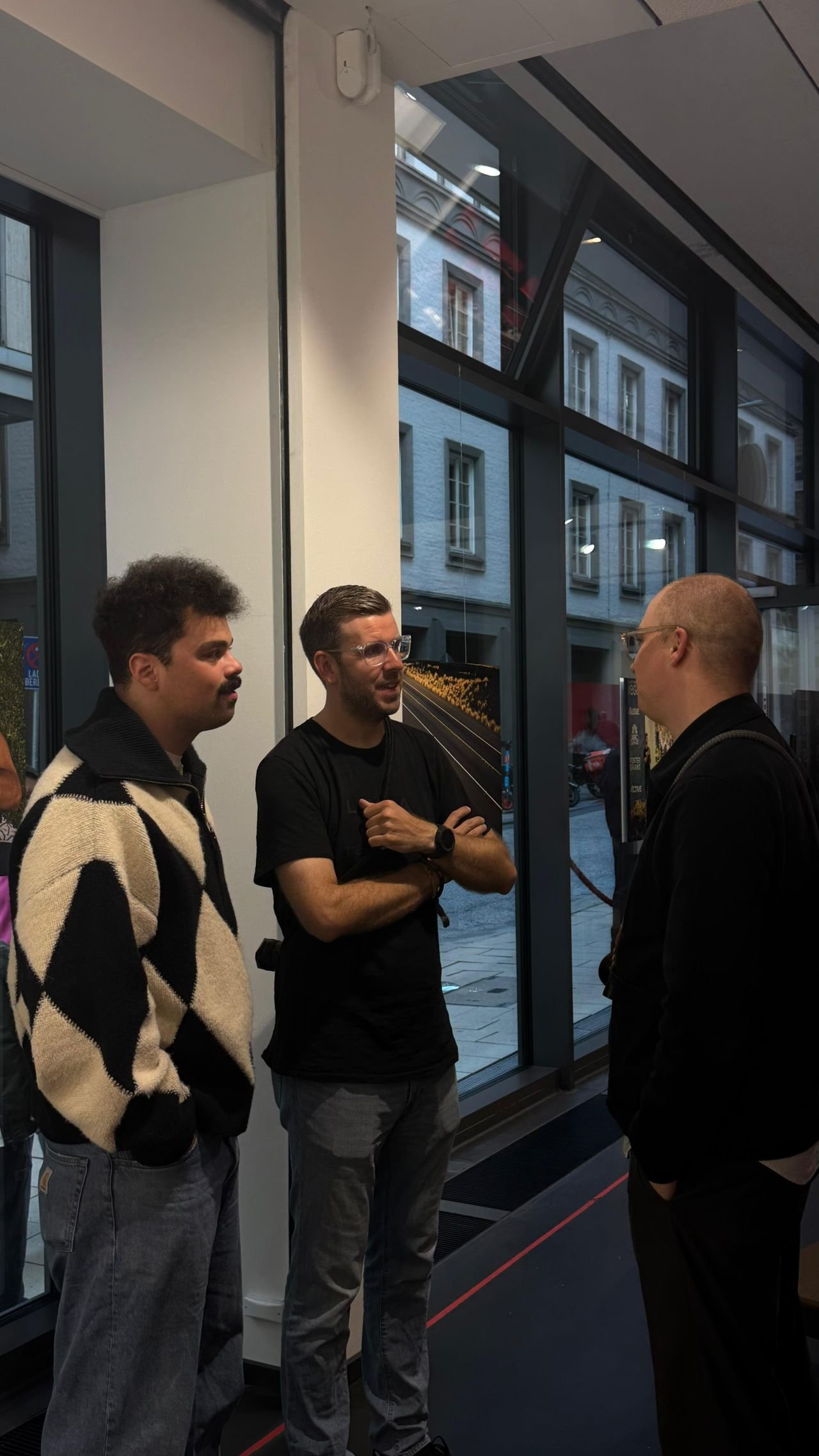 Drei Männer stehen im Gespräch am Fenster des Leica Stores, draußen die Straße bei Abendlicht