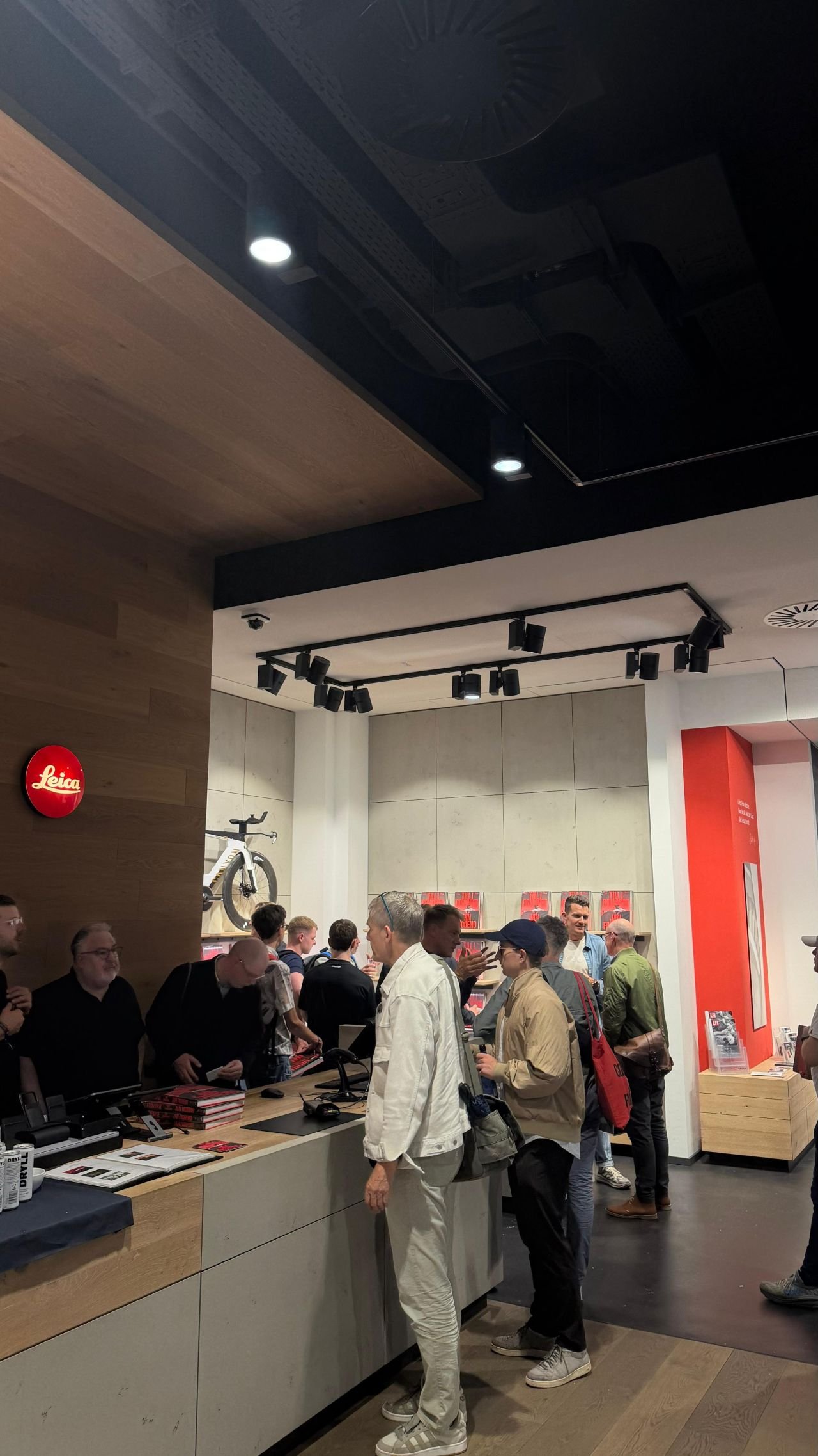 Blick in den Leica Store Hamburg mit rotem Leica-Logo an der Holzwand, Gästen am Verkaufstisch und Fotobüchern in den Regalen
