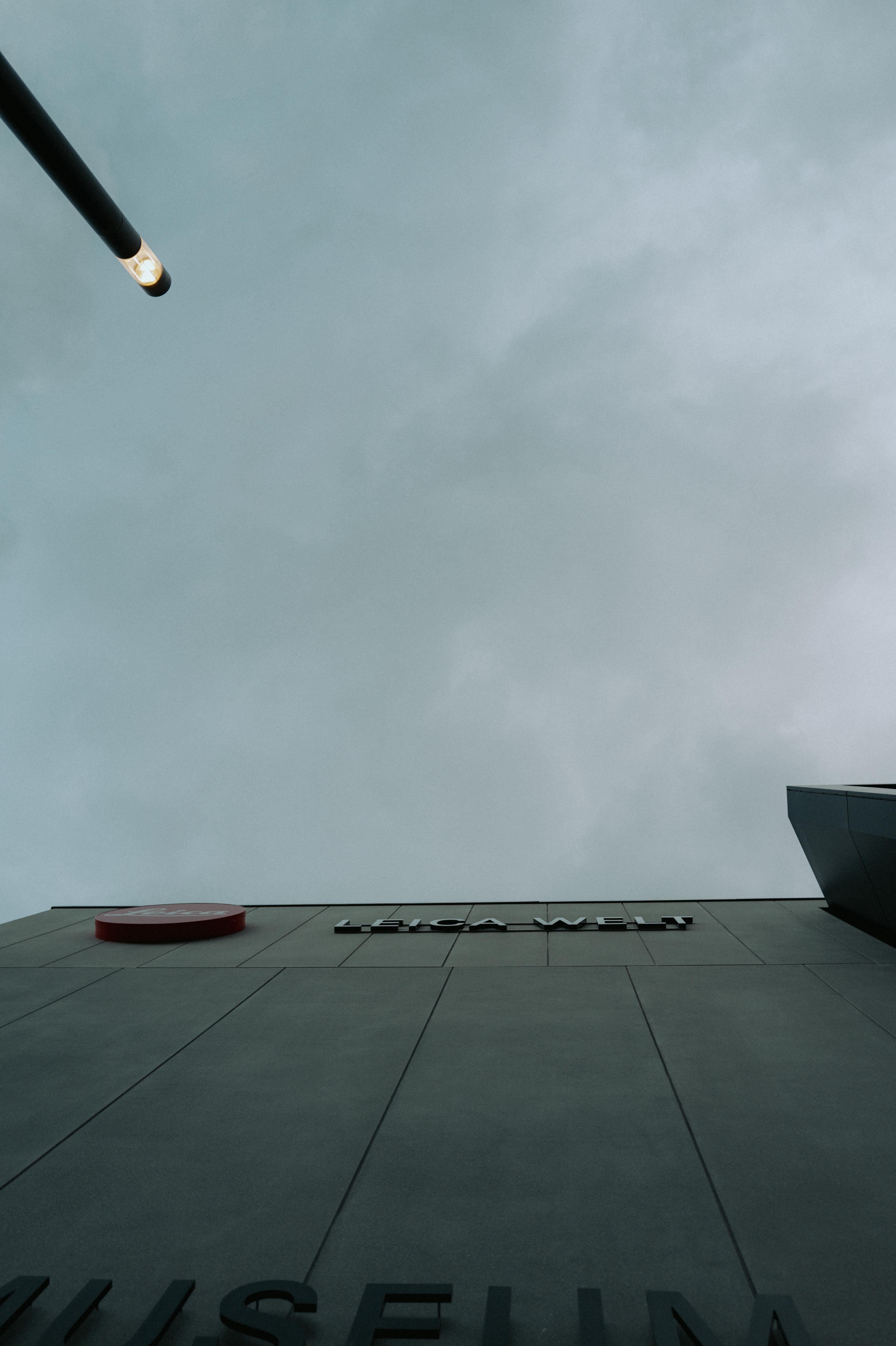 Blick nach oben an der Fassade der Leica Welt mit rotem Leica-Logo und Schriftzug vor bedecktem Himmel
