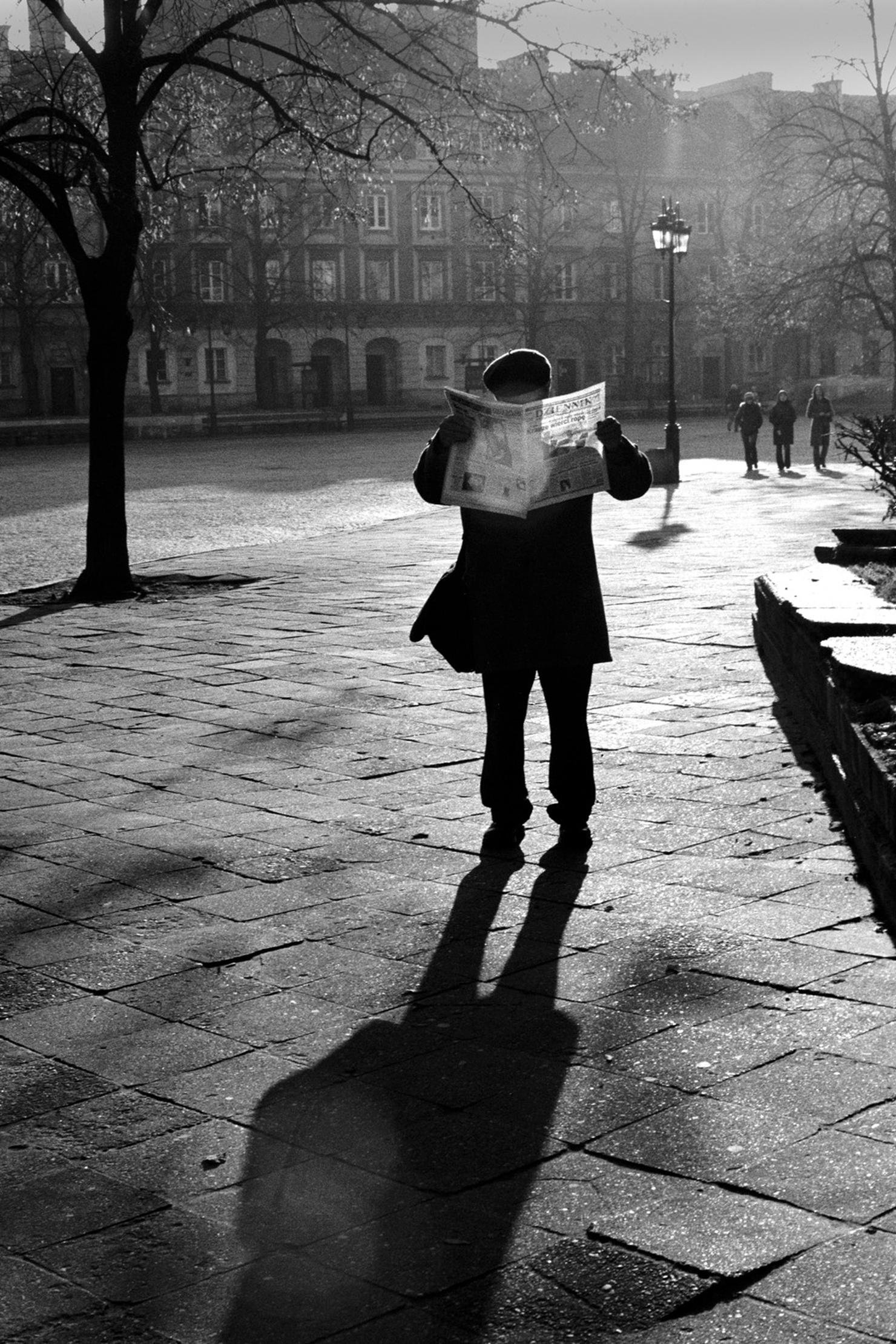 Schwarzweiß-Fotografie: ein Mann steht im Gegenlicht auf einem Platz in Warschau und liest Zeitung, sein Schatten fällt lang über das Pflaster