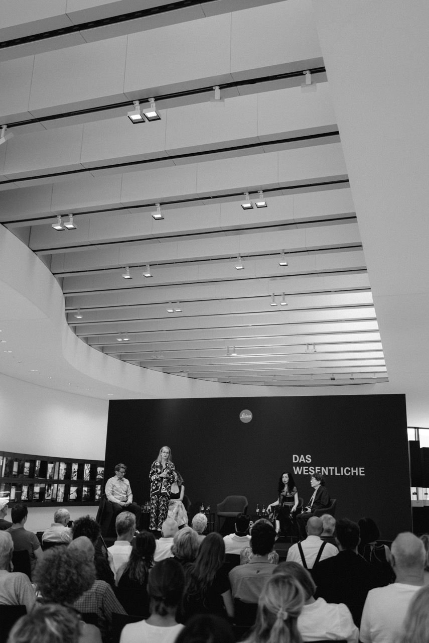 Podium mit vier Personen vor vollem Publikum, im Hintergrund eine dunkle Wand mit Leica-Logo und dem Schriftzug „Das Wesentliche“