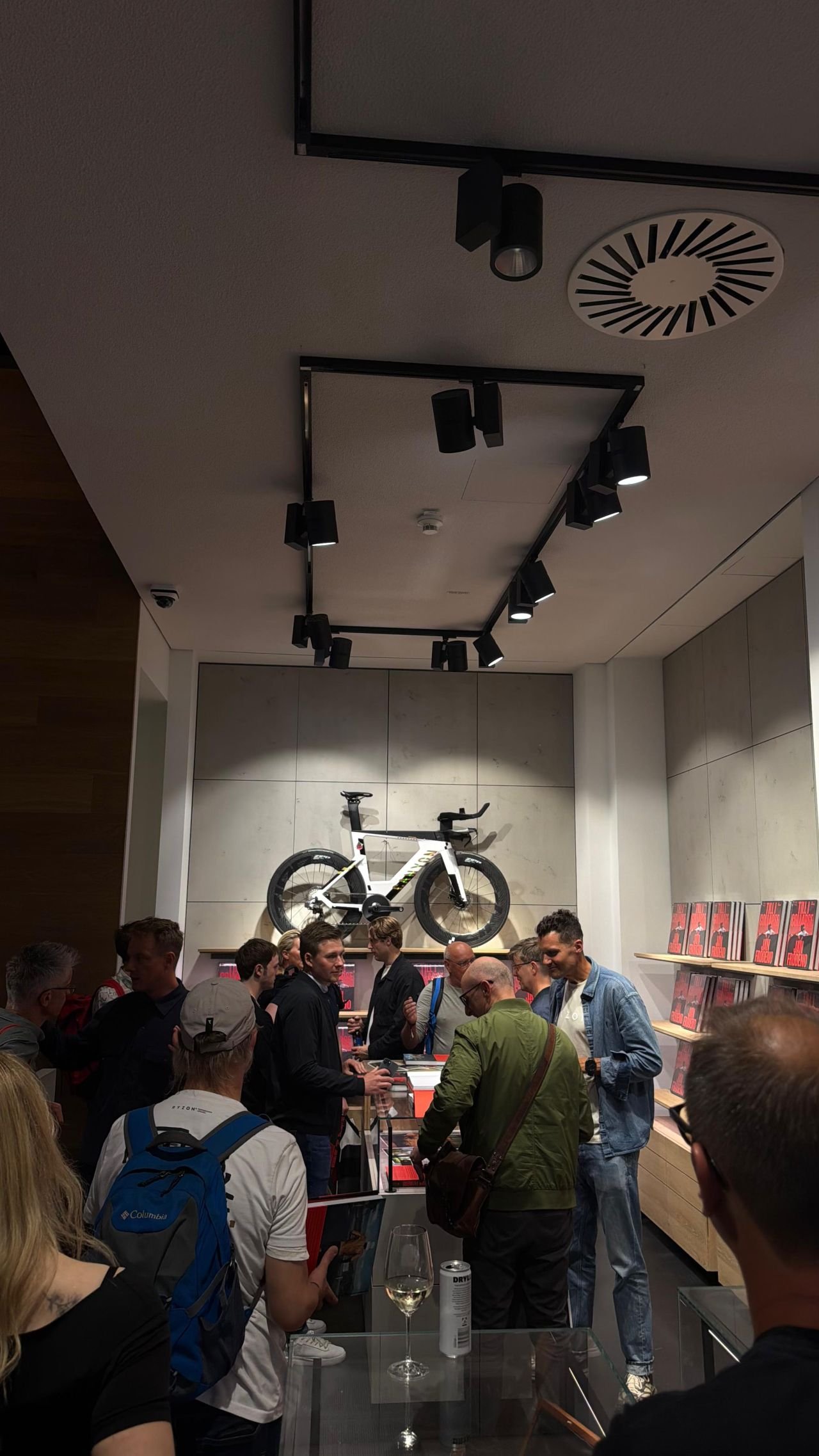 Zeitfahrrad an der Wand über einem Regal mit Fotobüchern, davor Gäste im Gespräch