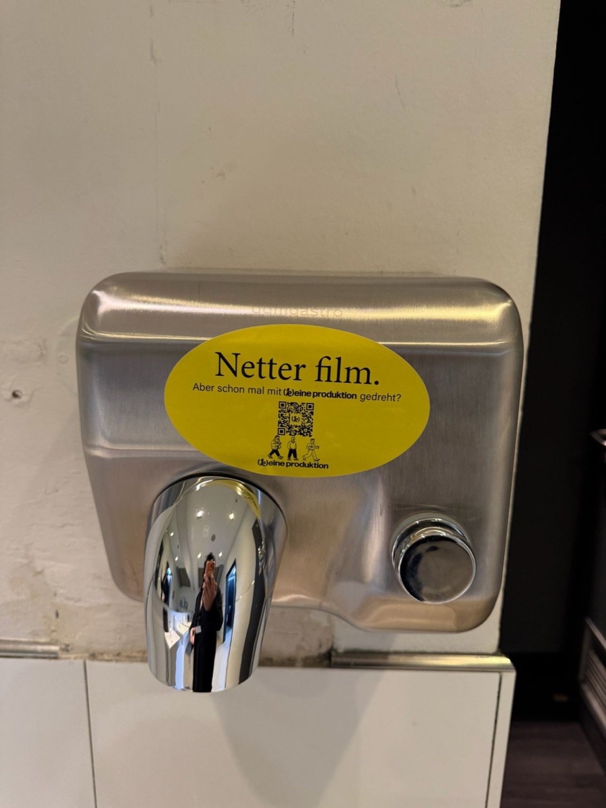 Sticker von [k]eine produktion zwischen anderen Aufklebern an einer Wand