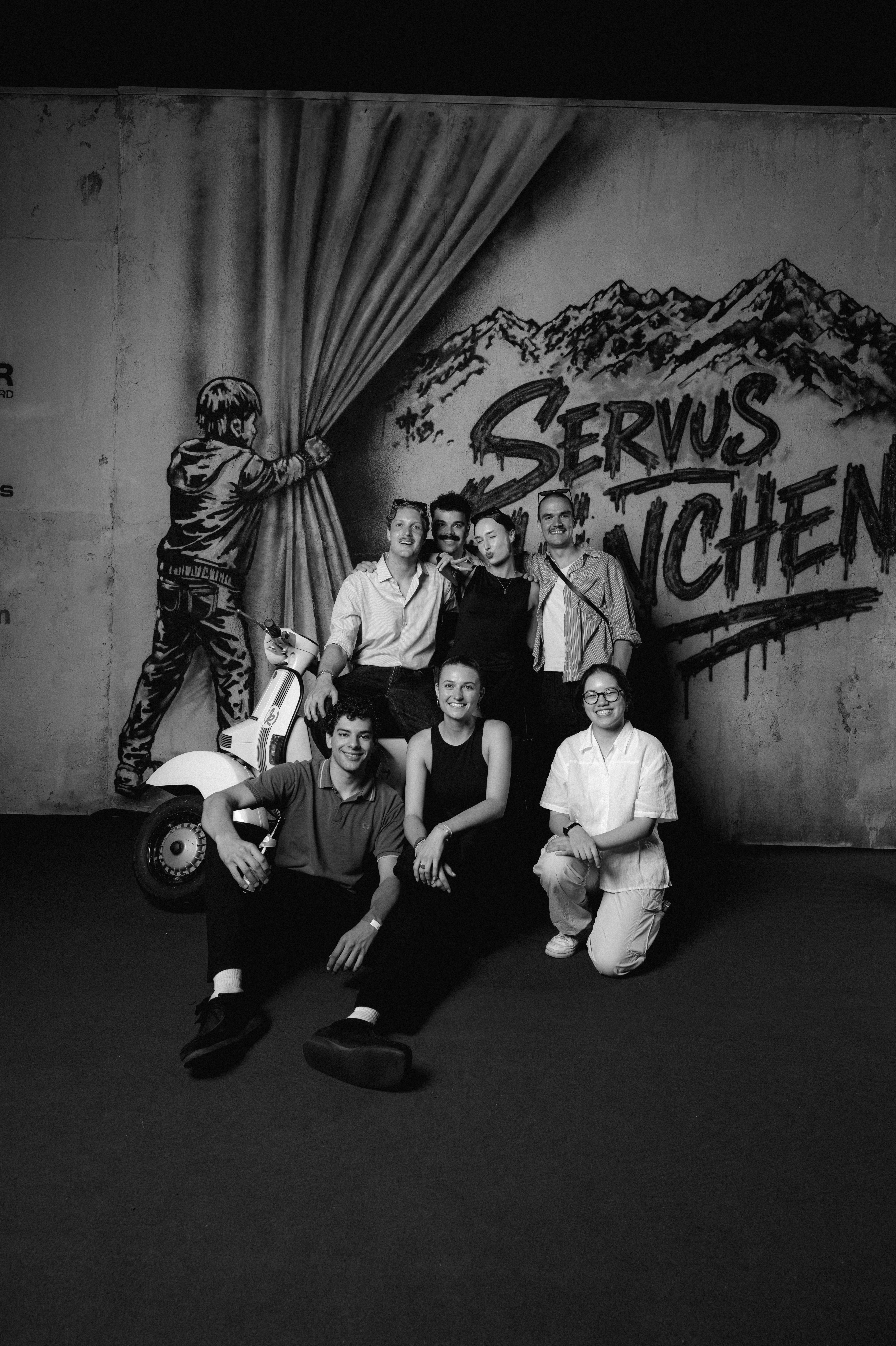 Gruppenfoto des Teams von [k]eine produktion vor der Servus München Wand mit der gebrandeten Vespa