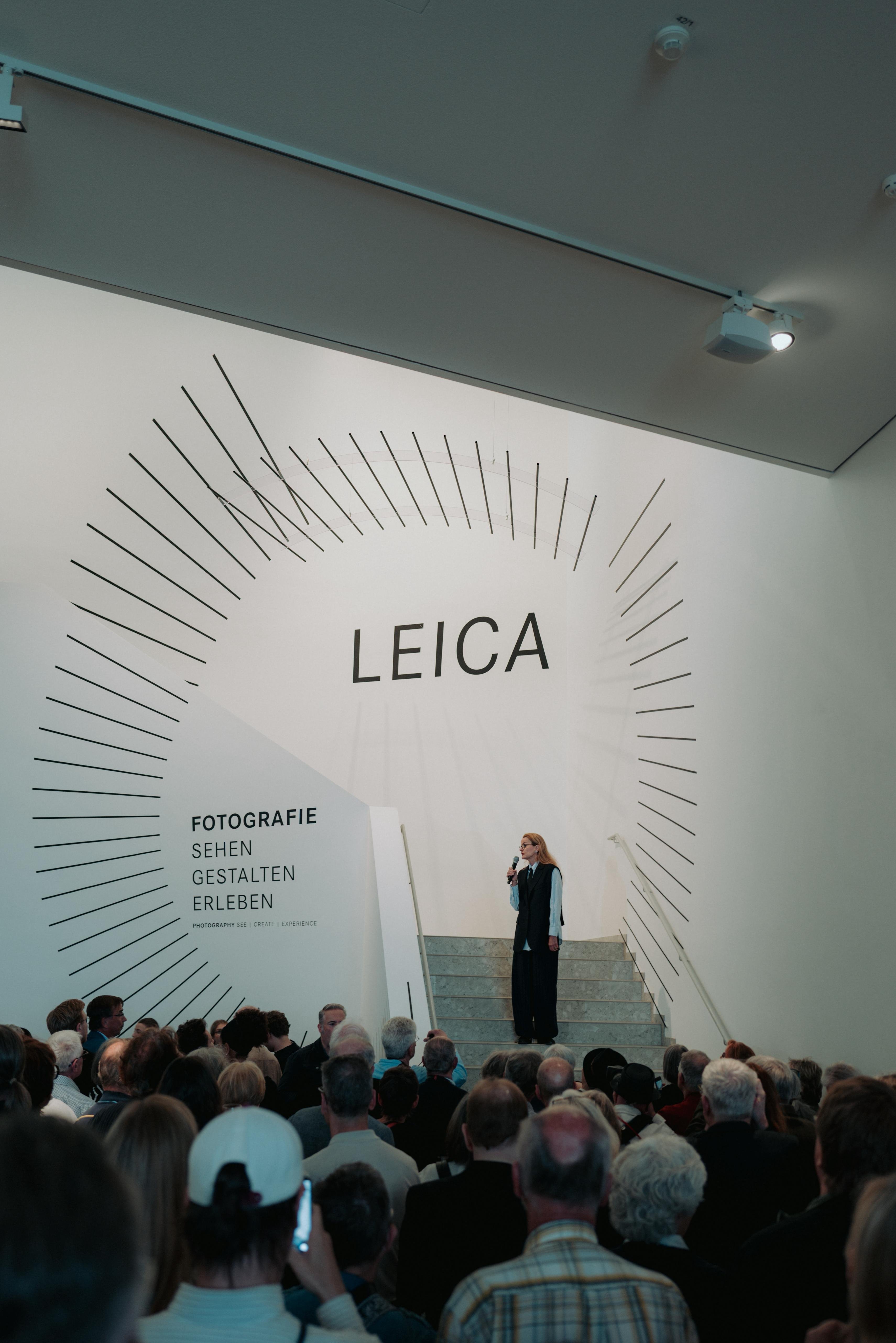 Eröffnungsrede zur Vernissage vor großem Publikum, dahinter ein riesiger LEICA-Schriftzug an der Wand