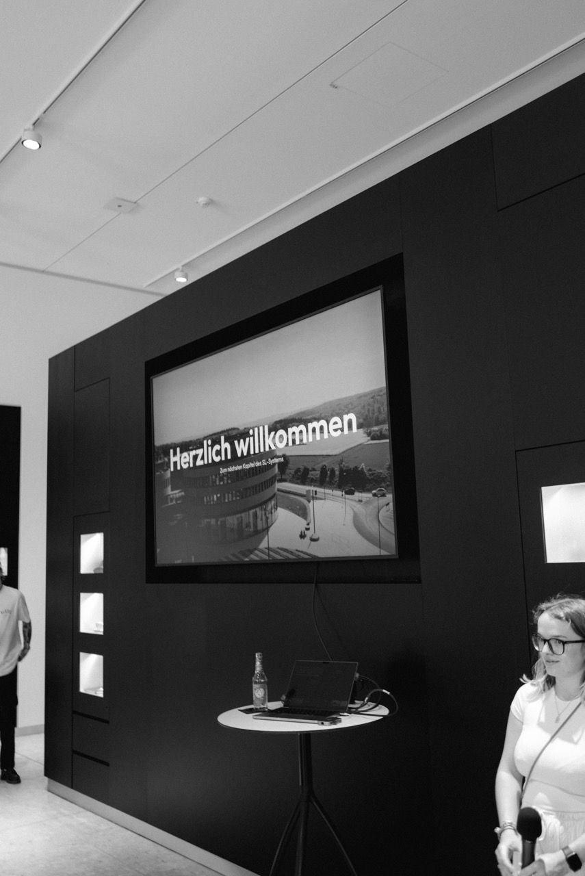 Bildschirm mit dem Schriftzug „Herzlich willkommen. Zum nächsten Kapitel des SL-Systems“ in einem abgedunkelten Ausstellungsraum